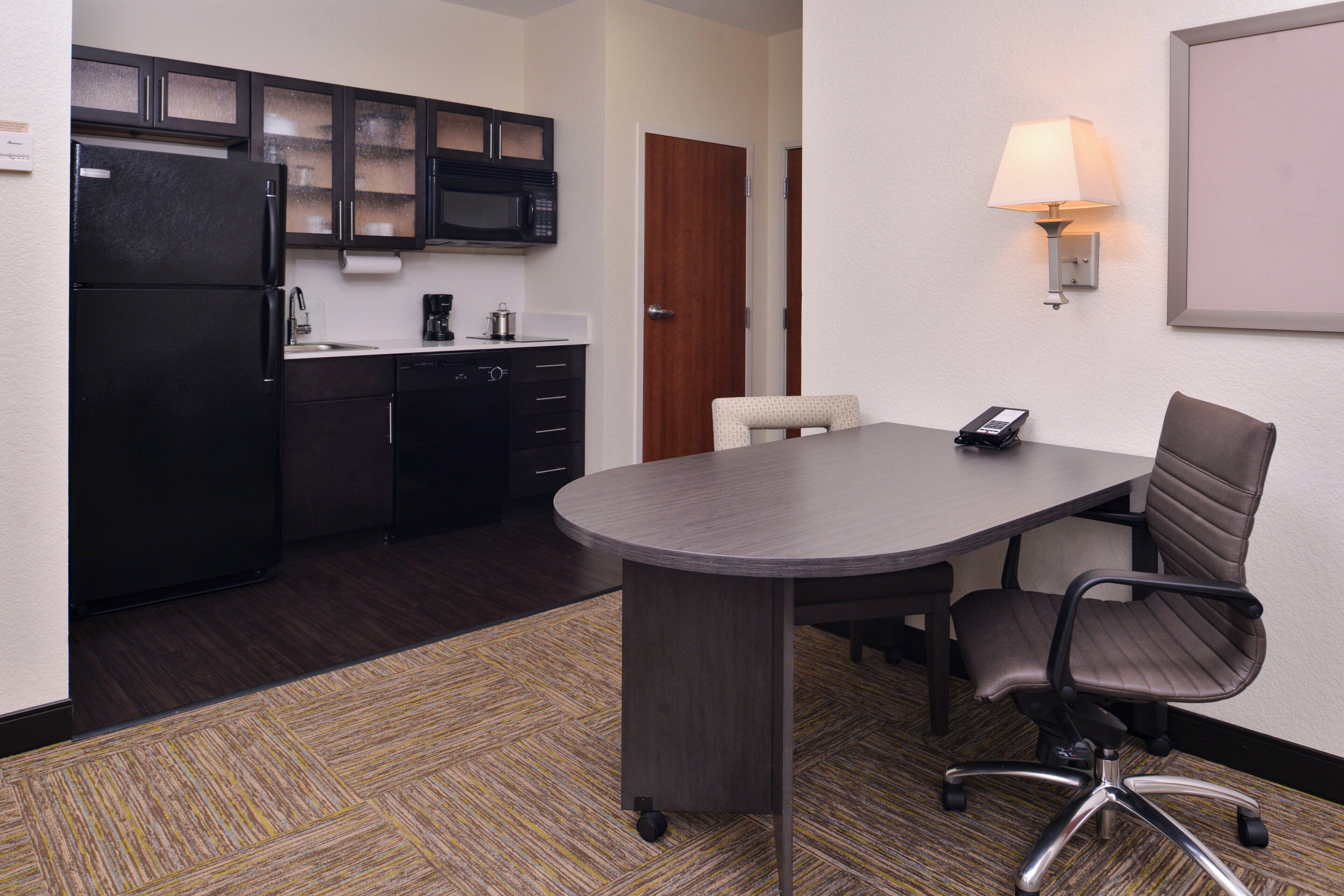 A hotel room with a kitchenette, a conference table, a single office chair, and a lamp.