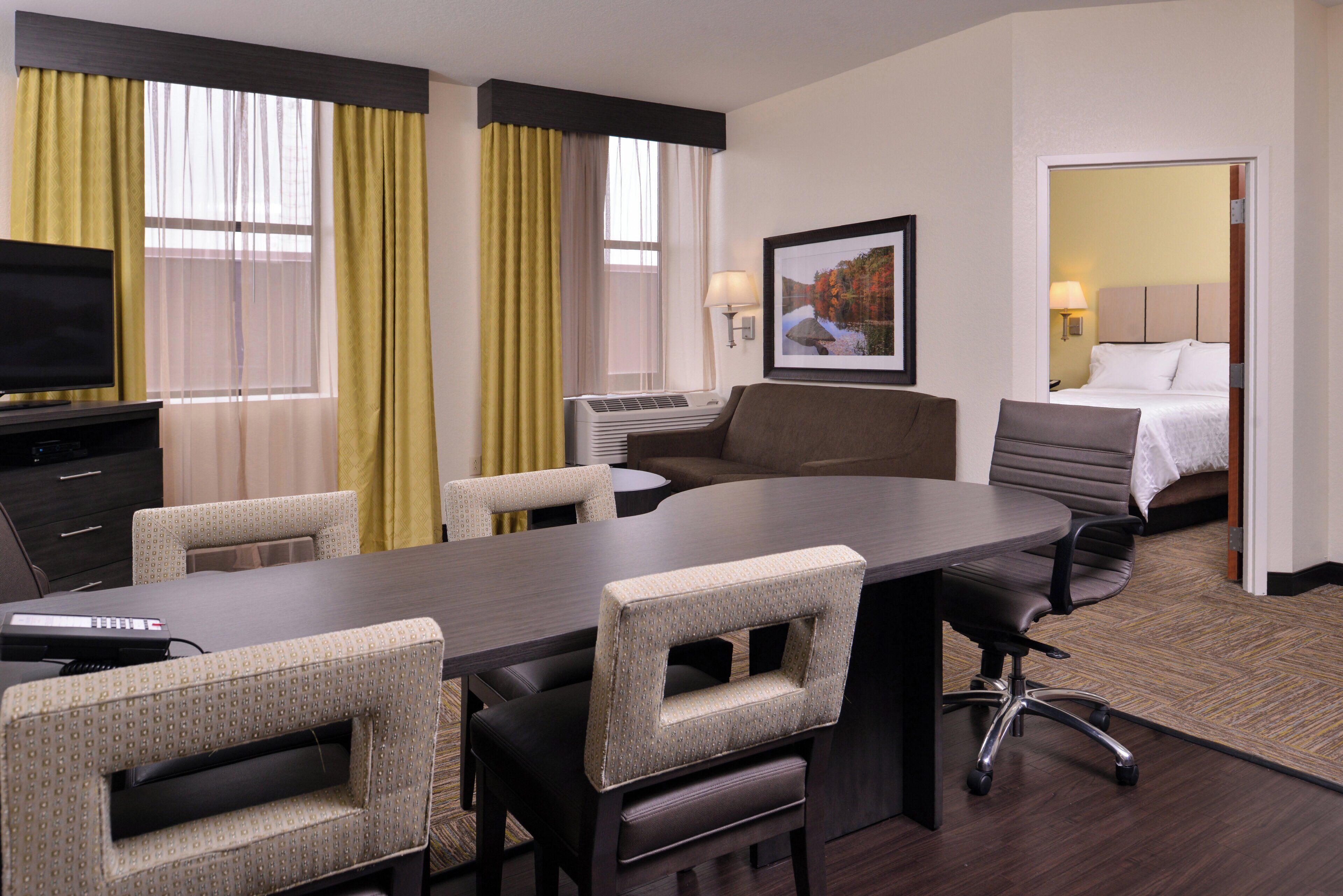 A hotel room with a conference table, chairs, a bed, a TV, and a painting on the wall.