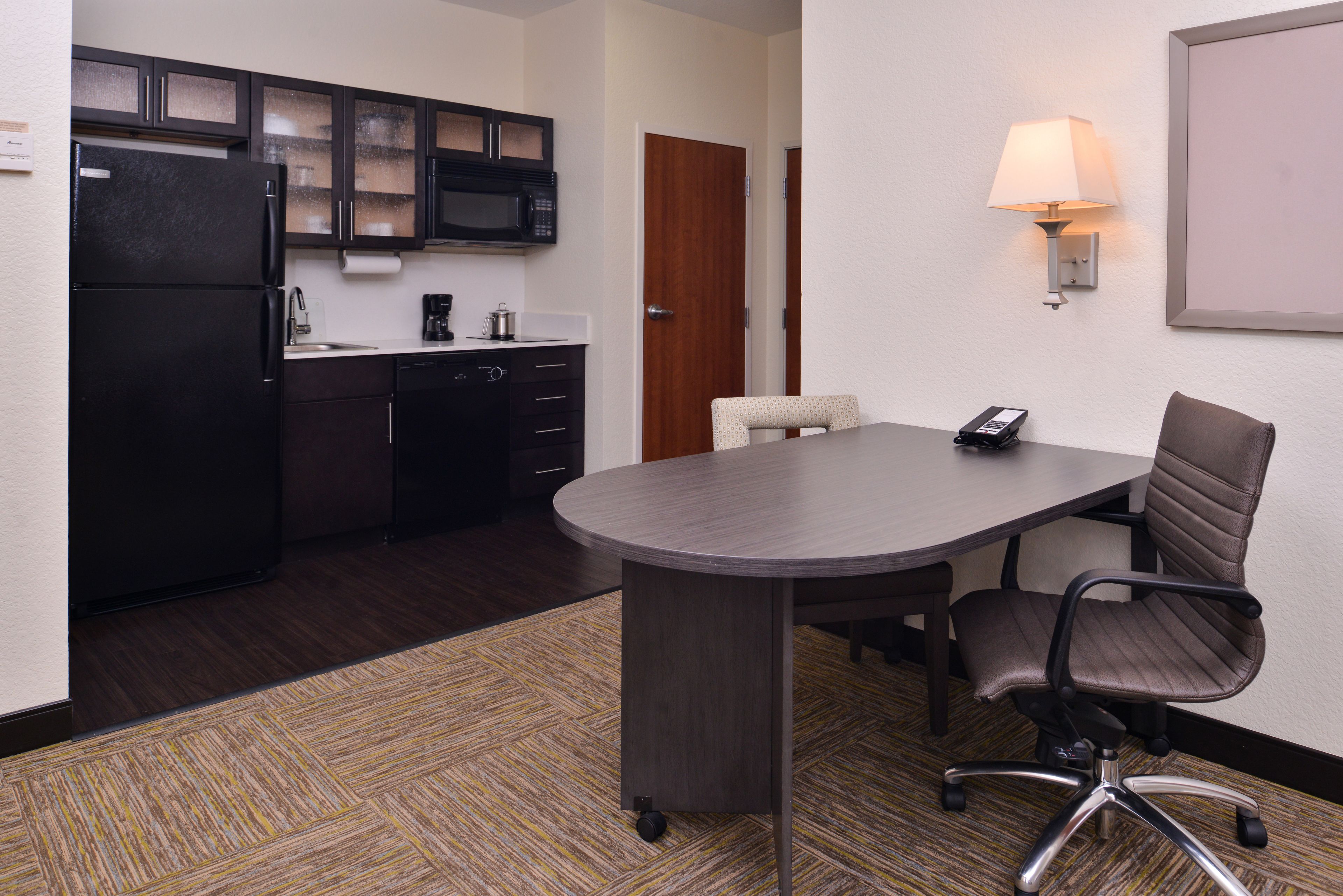 A conference room with a dark wooden table, a brown office chair, a black refrigerator, and a microwave in the adjacent kitchen area.