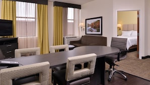 A hotel room with a large conference table, four chairs, a TV, an air conditioning unit, a bed, and a framed picture on the wall.