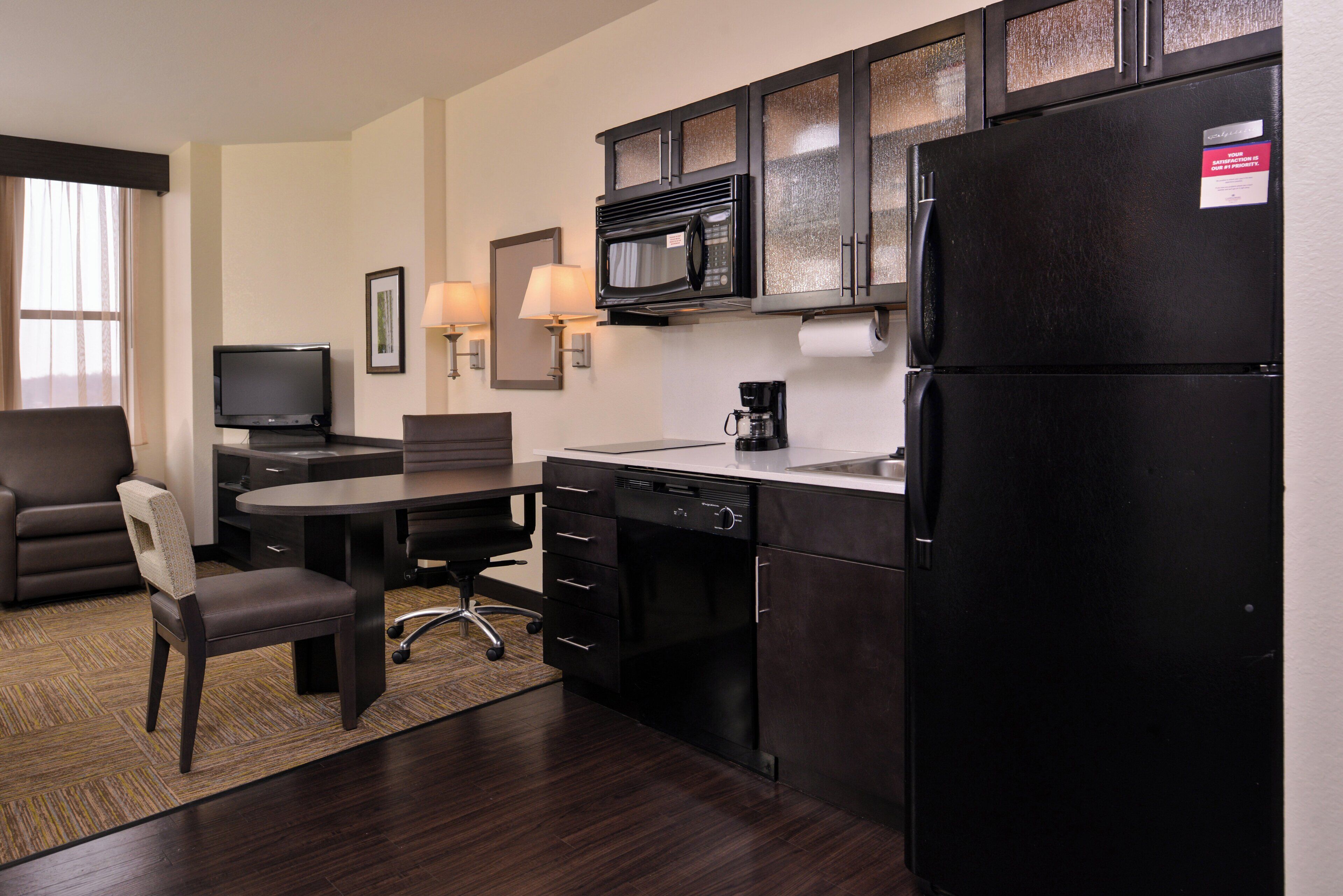 A compact hotel room with a kitchenette, a dining area, a flat-screen TV, and a seating area with a sofa and a chair.
