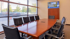 A conference room with a long wooden table, six black leather chairs, and a large window with blinds.