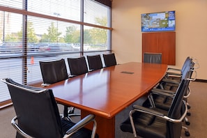 Een vergaderzaal met een lange houten tafel, zes zwarte lederen stoelen en een groot raam met zonwering.