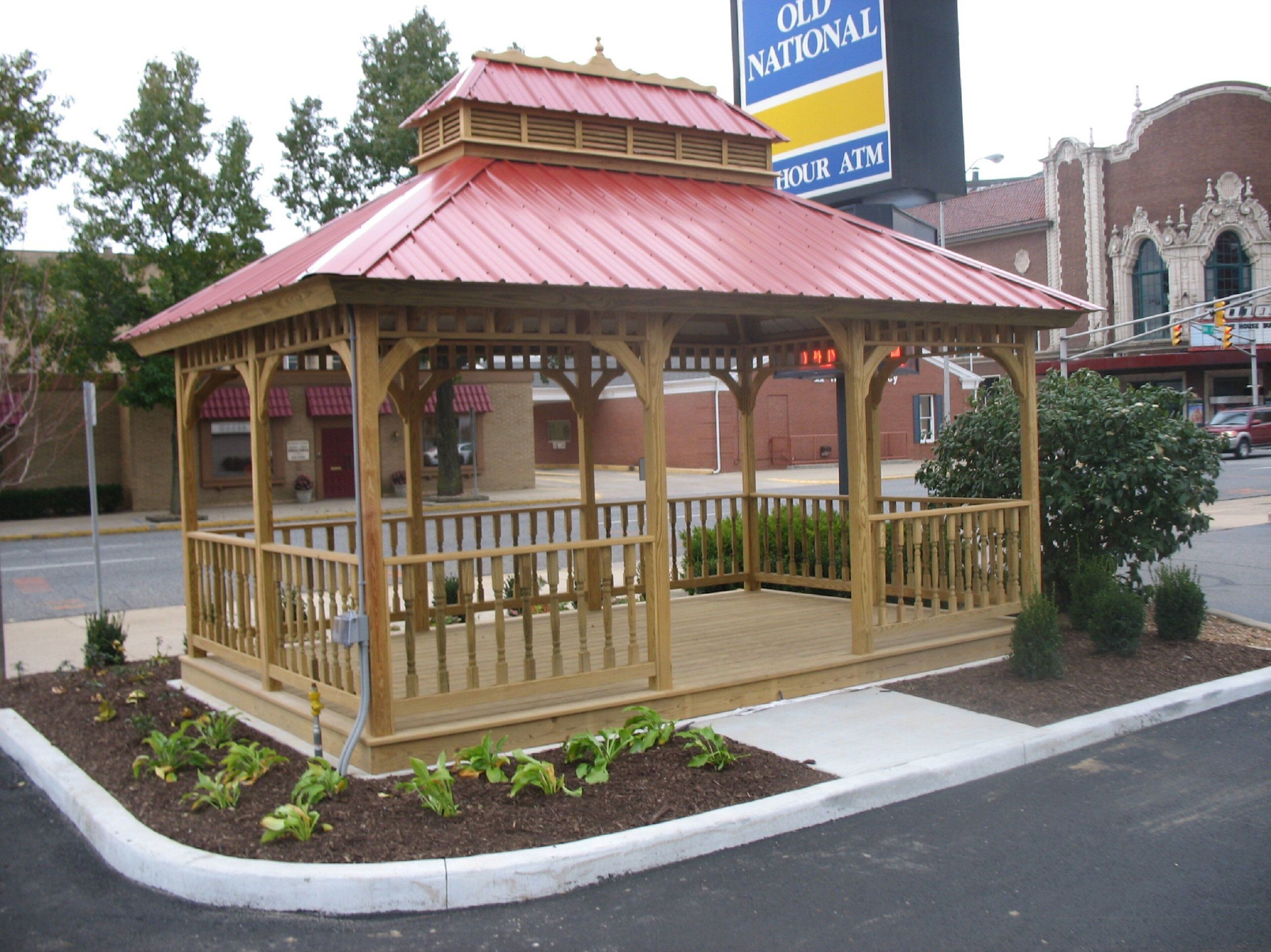 A gazebo with a red roof and wooden railings, surrounded by a landscaped garden with small plants.
