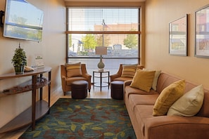 A hotel room with a sofa, two armchairs, a small table, a potted plant, and a window with blinds.