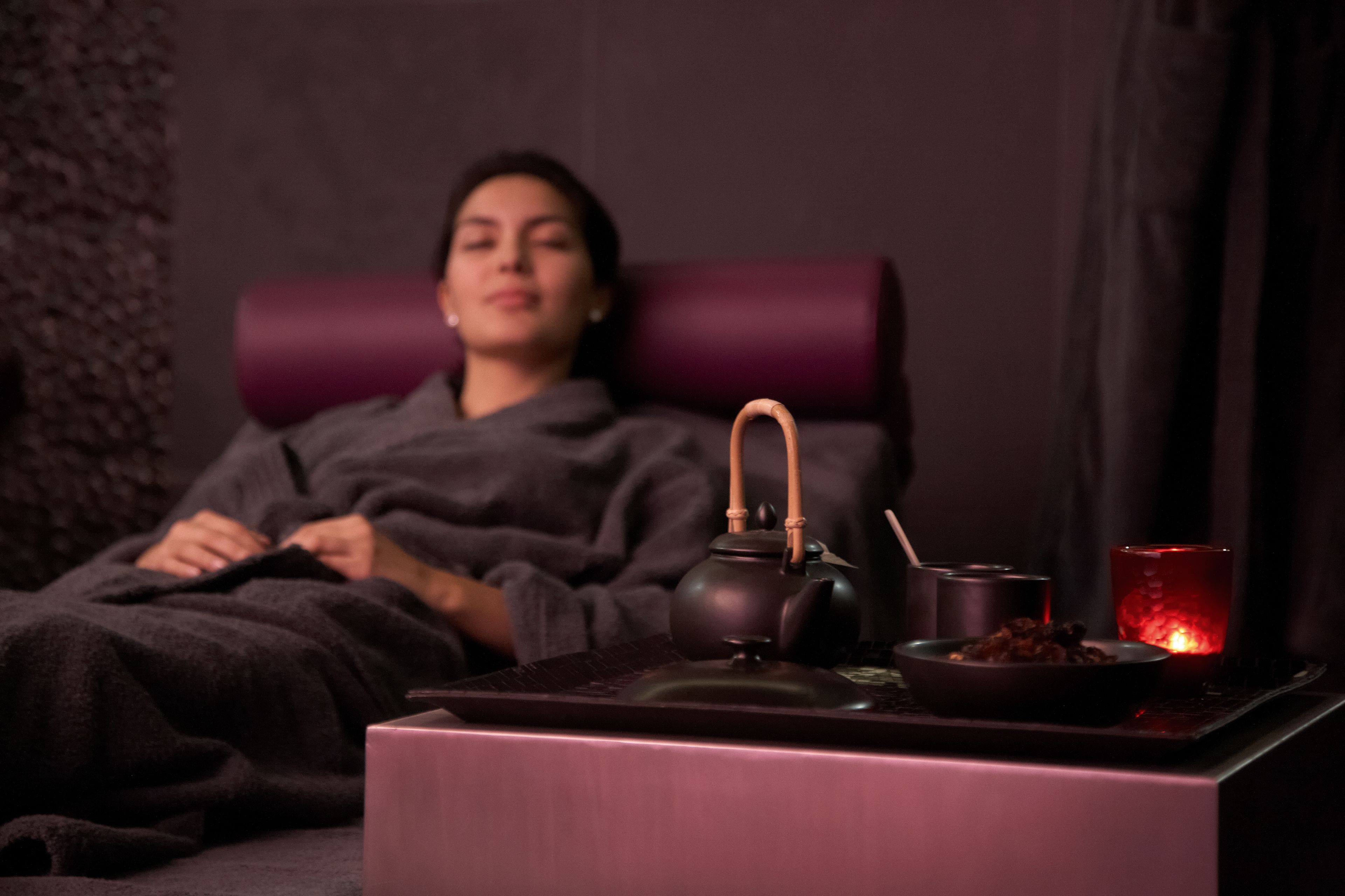 A person lying in a chair with a teapot, a bowl, and a candle on a table.