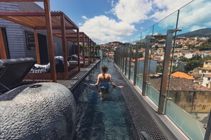 Una piscina infinita en la azotea con barandilla de vidrio, sillas de descanso y vistas a un paisaje urbano.
