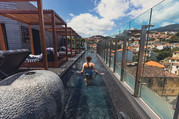 Una piscina infinita en la azotea con barandilla de vidrio, sillas de descanso y vistas a un paisaje urbano.
