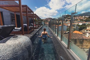 A rooftop infinity pool with a glass balustrade, lounge chairs, and a view of a cityscape.