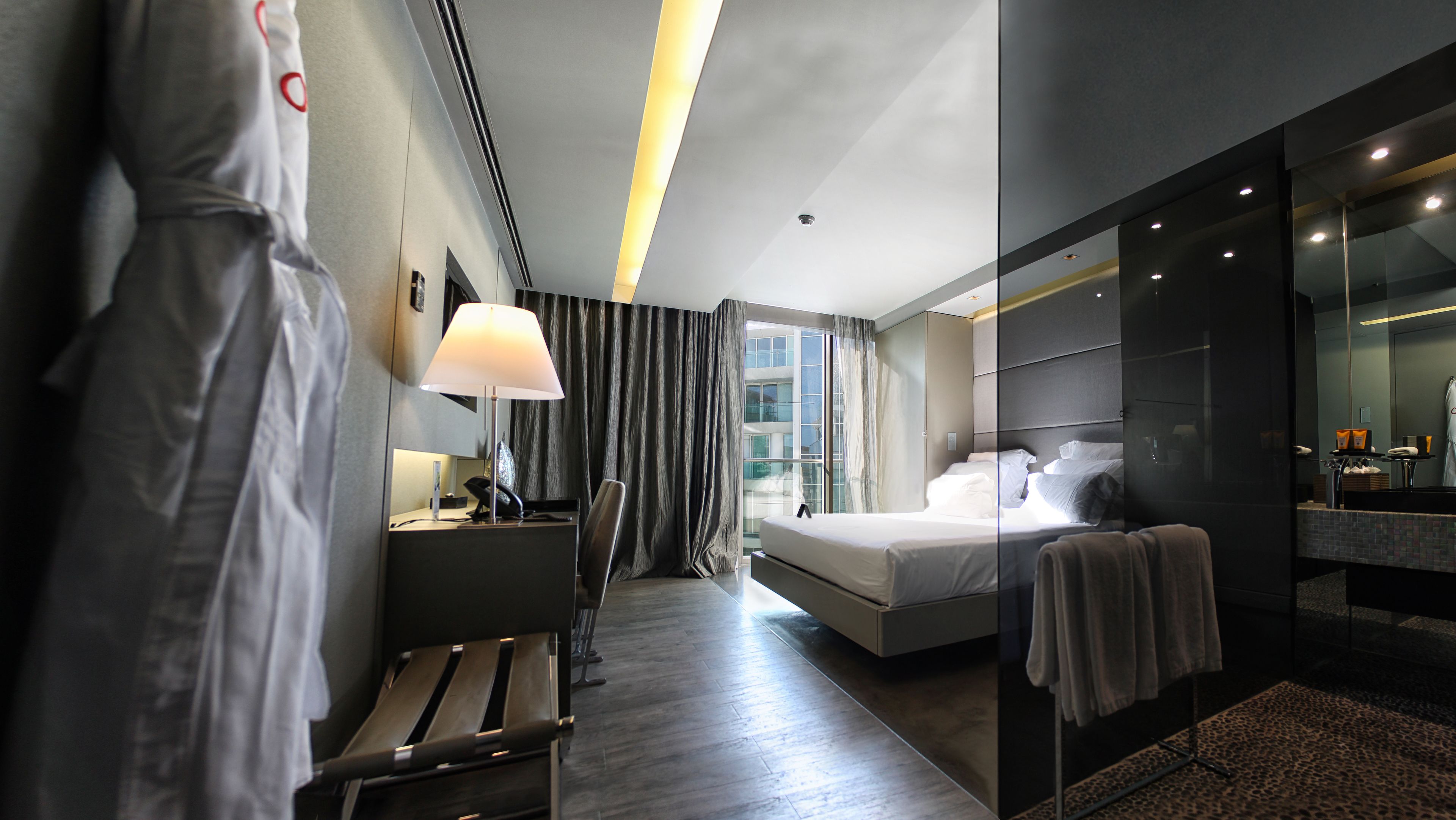 A modern hotel room with a large bed, a desk with a lamp, a chair, and a bathroom with a mirror and sink.