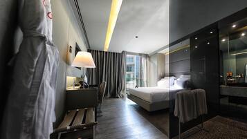 A modern hotel room with a large bed, a desk with a lamp, a chair, and a bathroom with a mirror and sink.