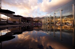 A modern rooftop pool with glass railings and lounge chairs, reflecting a cityscape and mountains in the background. - The Vine (Funchal)