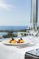A table with a plate of colorful desserts and two empty wine glasses, set against a backdrop of a clear sky and a distant view of the sea.