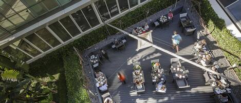 An outdoor dining area with tables and chairs, surrounded by greenery and a modern building with large windows.