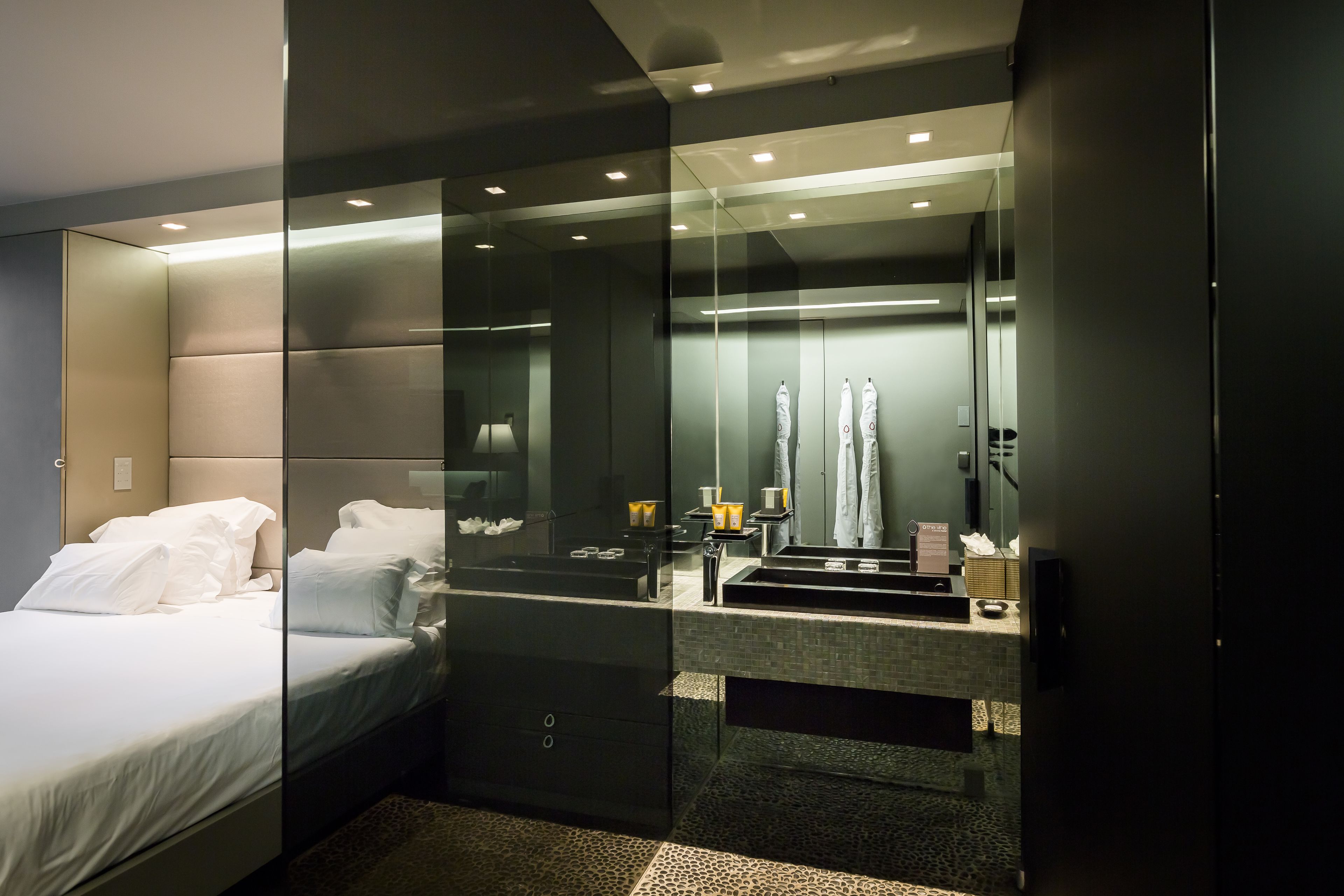 A modern bedroom with a glass partition, a bed with white linens, a bedside table, and a mirror reflecting the room.