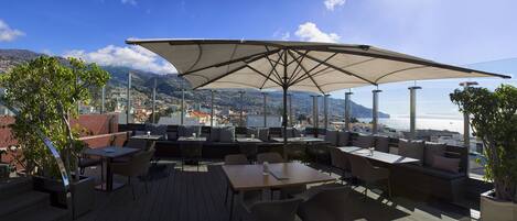 An outdoor seating area with tables and chairs, a large umbrella, and a view of the city and sea.