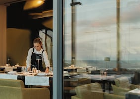 Un restaurant avec des tables dressées, un serveur s’occupant d’une table, et une vue à travers le mur de verre.