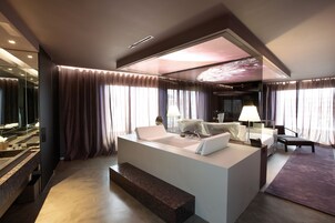 A modern hotel room with a large bed, a desk, a chair, and a glass-enclosed bathroom.
