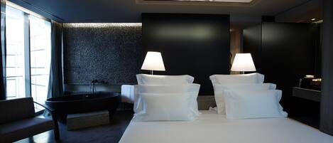 A modern hotel room with a large bed, two bedside lamps, a bathtub, and a view of the city through large windows.