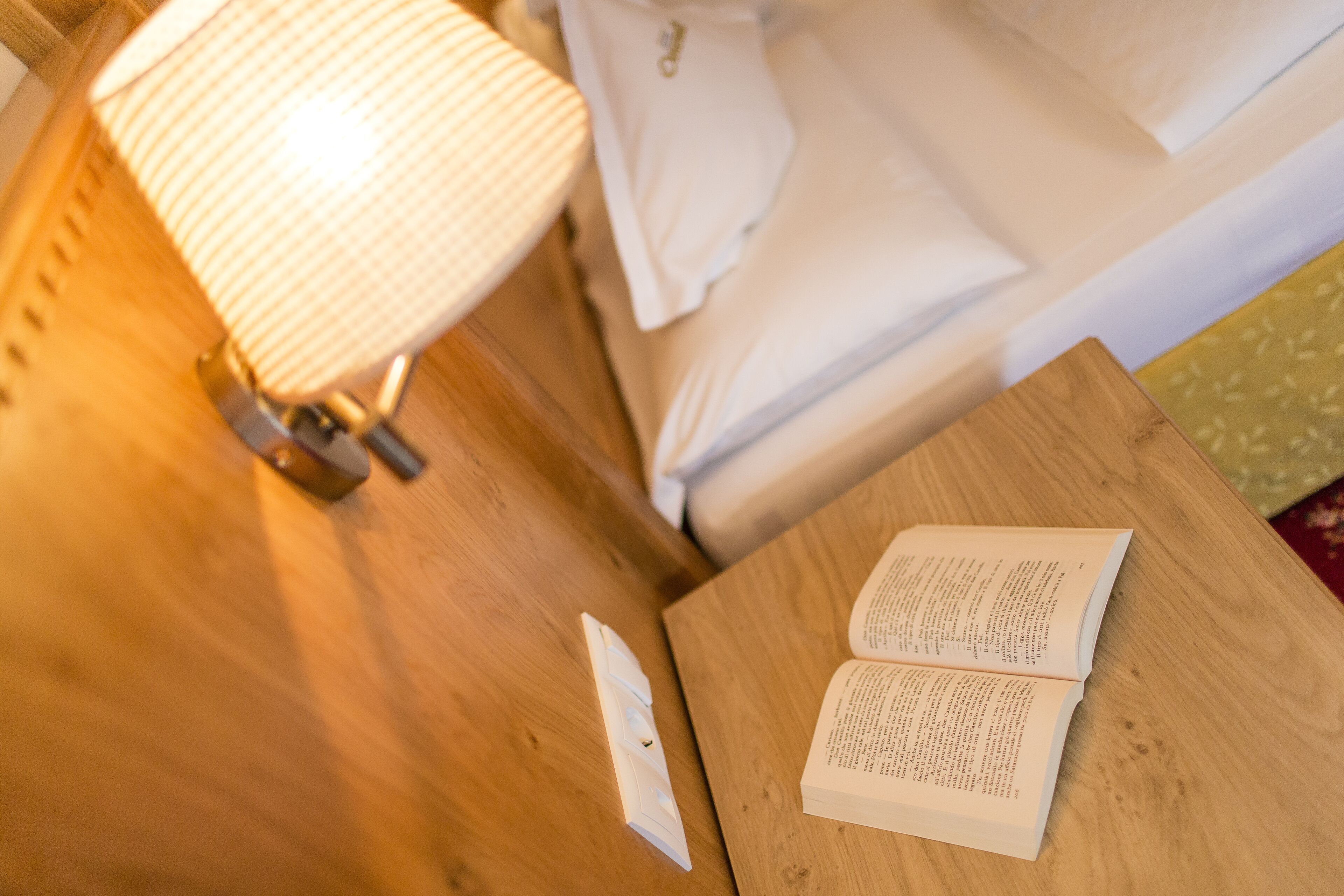 A wooden bedside table with an open book, a lamp, and a bed in the background.