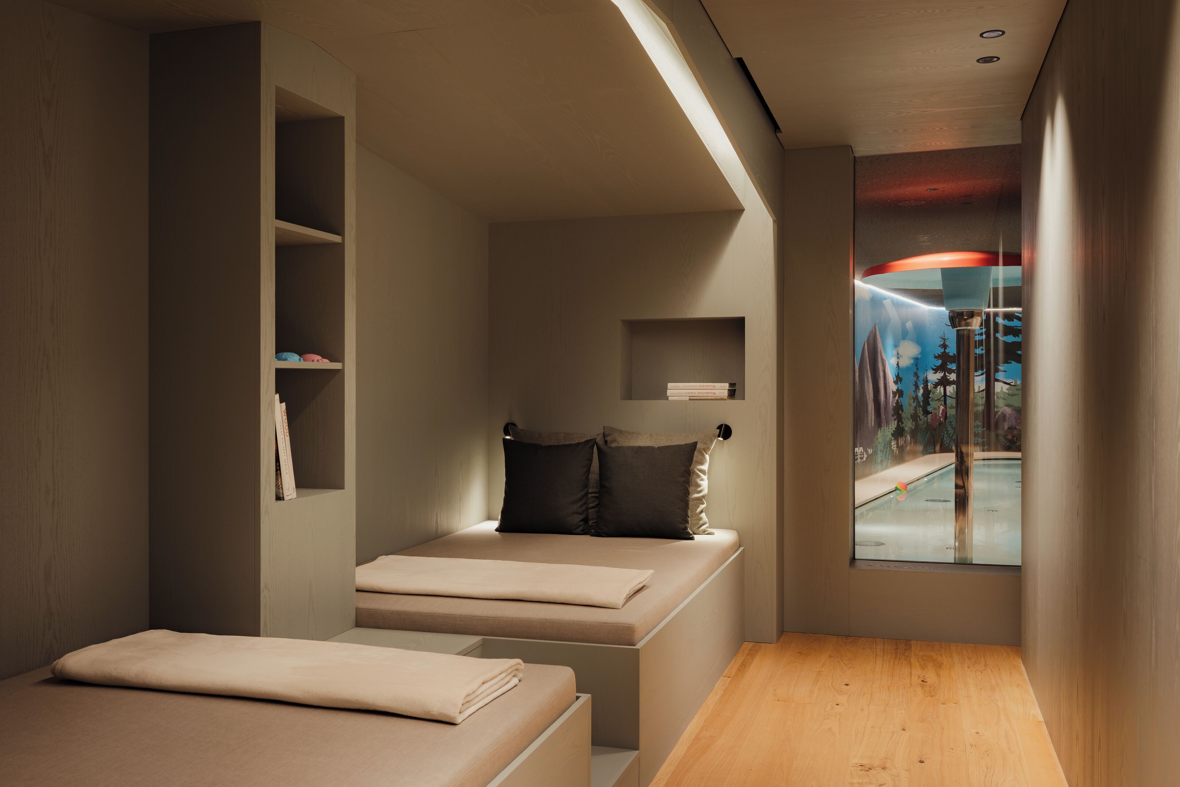 A modern, compact bedroom with a built-in bed, a small seating area, and a window with a view of trees and a swimming pool.