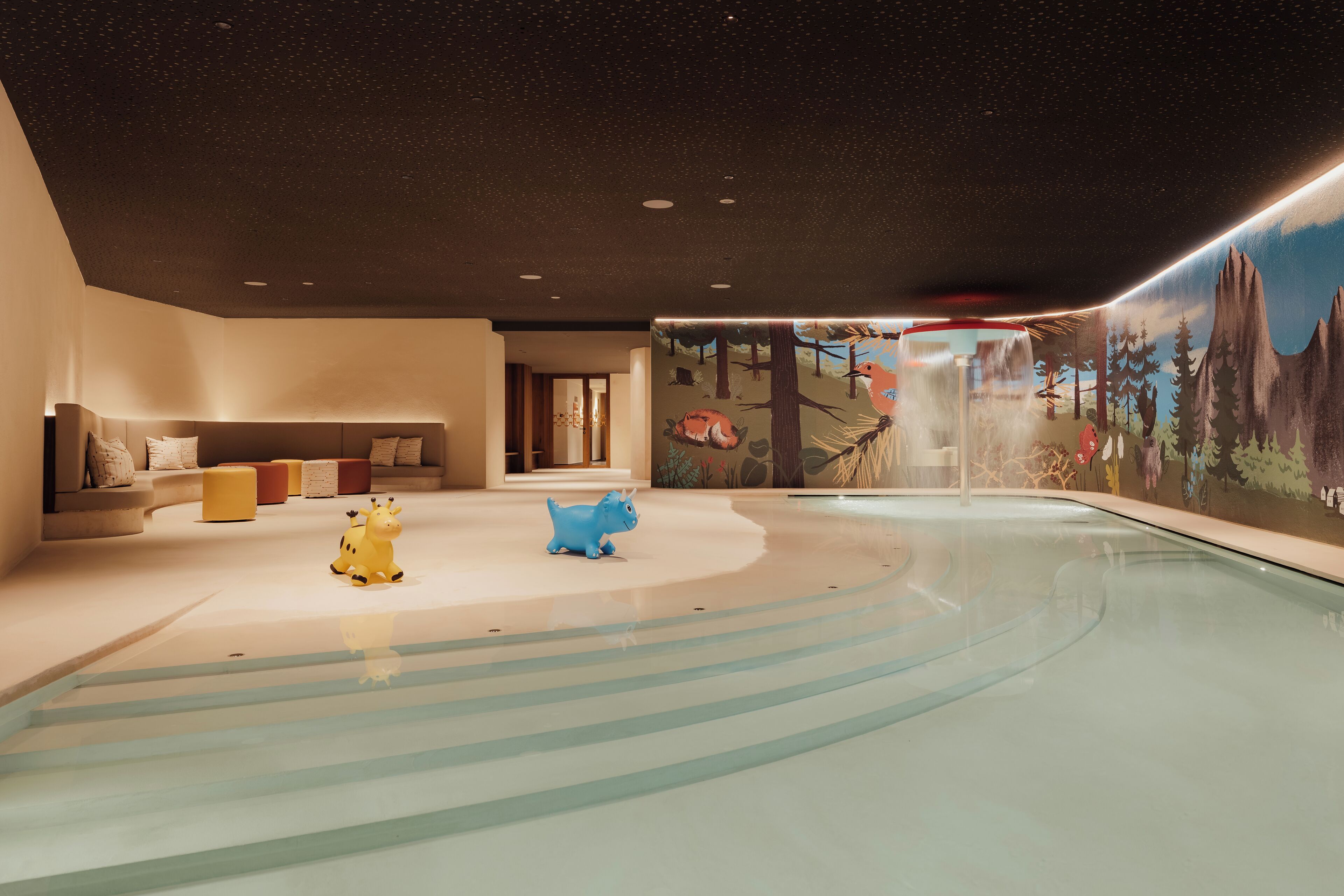 A modern indoor pool area with a starry ceiling, a mural of a forest, and a seating area with cushions.