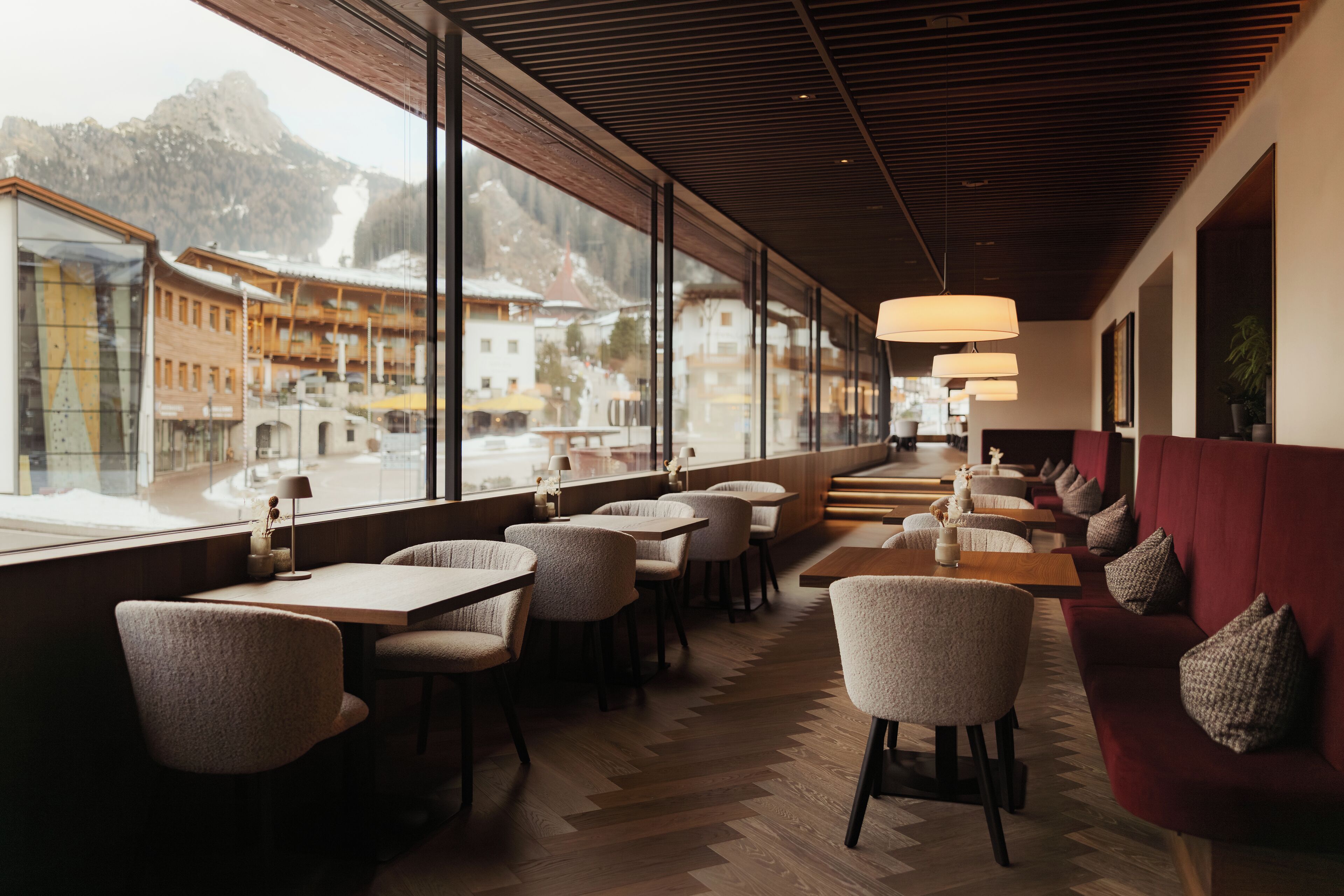A modern interior with wooden flooring, tables, chairs, and a red sofa. Large windows offer a view of a snowy mountain landscape.
