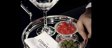 Una bandeja con una copa de martini, una servilleta con el texto 'The Oswald Grand Hotel' y dos cuencos de aceitunas.
