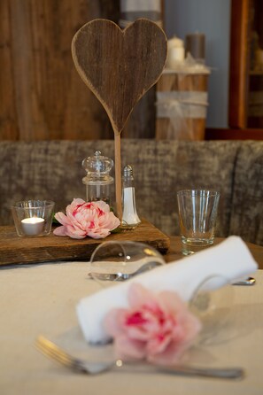 A table setting with a wooden heart-shaped centerpiece, a glass candle holder, a clear glass, and a pink flower. - Genuss-Boutique B&B Wienerhof (Trins)