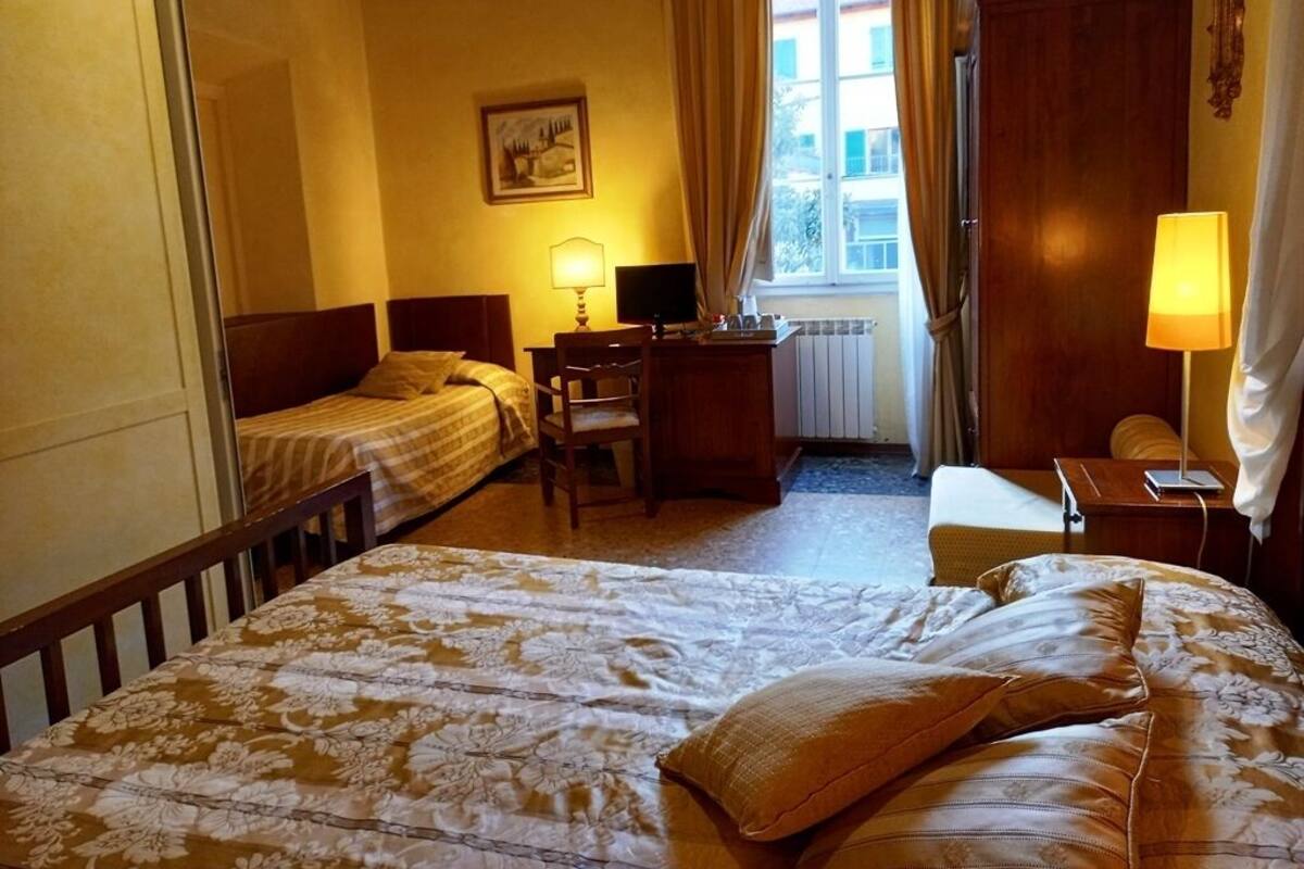 Une chambre d’hôtel avec deux lits simples, un bureau avec une chaise, une petite table avec une lampe et une fenêtre avec des rideaux.