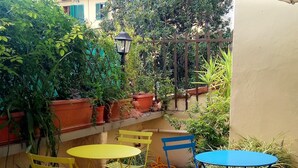 Un patio al aire libre con varias mesas y sillas, rodeado de macetas con plantas y una sombrilla grande que proporciona sombra.