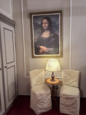 Ein Zimmer mit einem Gemälde der Mona Lisa an der Wand, zwei Stühle mit einem Tisch dazwischen und einer Tür links.