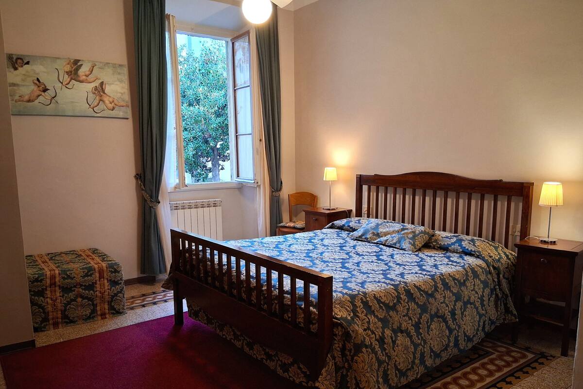 Une chambre à coucher comprenant un lit, une table de chevet, une lampe, une fenêtre avec des rideaux, un radiateur et une petite table avec une chaise.