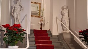 Una escalera con alfombra roja, estatuas blancas y plantas en macetas.
