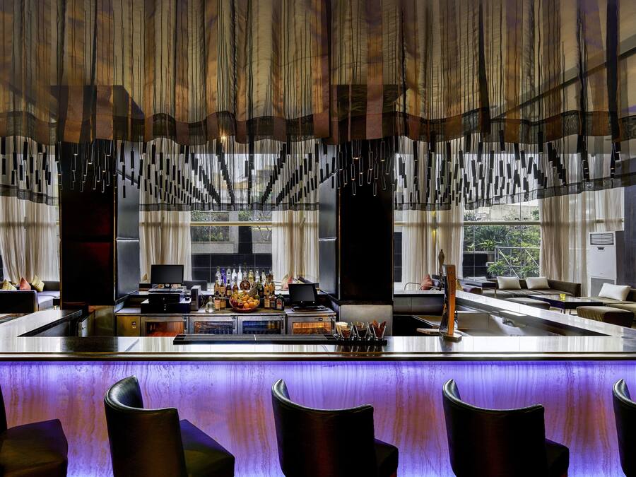 Un área de bar moderna con una barra alargada, taburetes y vistas al exterior a través de amplios ventanales.