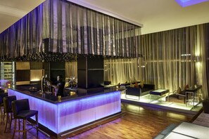 A modern bar area with a bar counter, stools, and a seating area with sofas and a coffee table. - Taj Club House (Chennai)