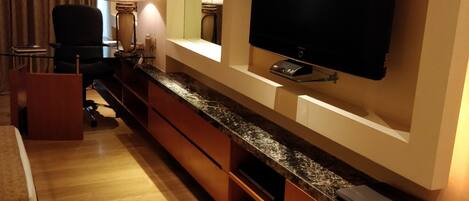 Una habitación de hotel moderna con televisión de pantalla plana montada en la pared, suelo de madera, un escritorio con lámpara, una silla y un gran ventanal con cortinas.