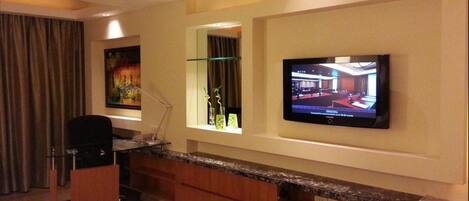 Habitación de hotel con cama, escritorio, televisor montado en la pared, ventana con cortinas y un cuadro colgado.