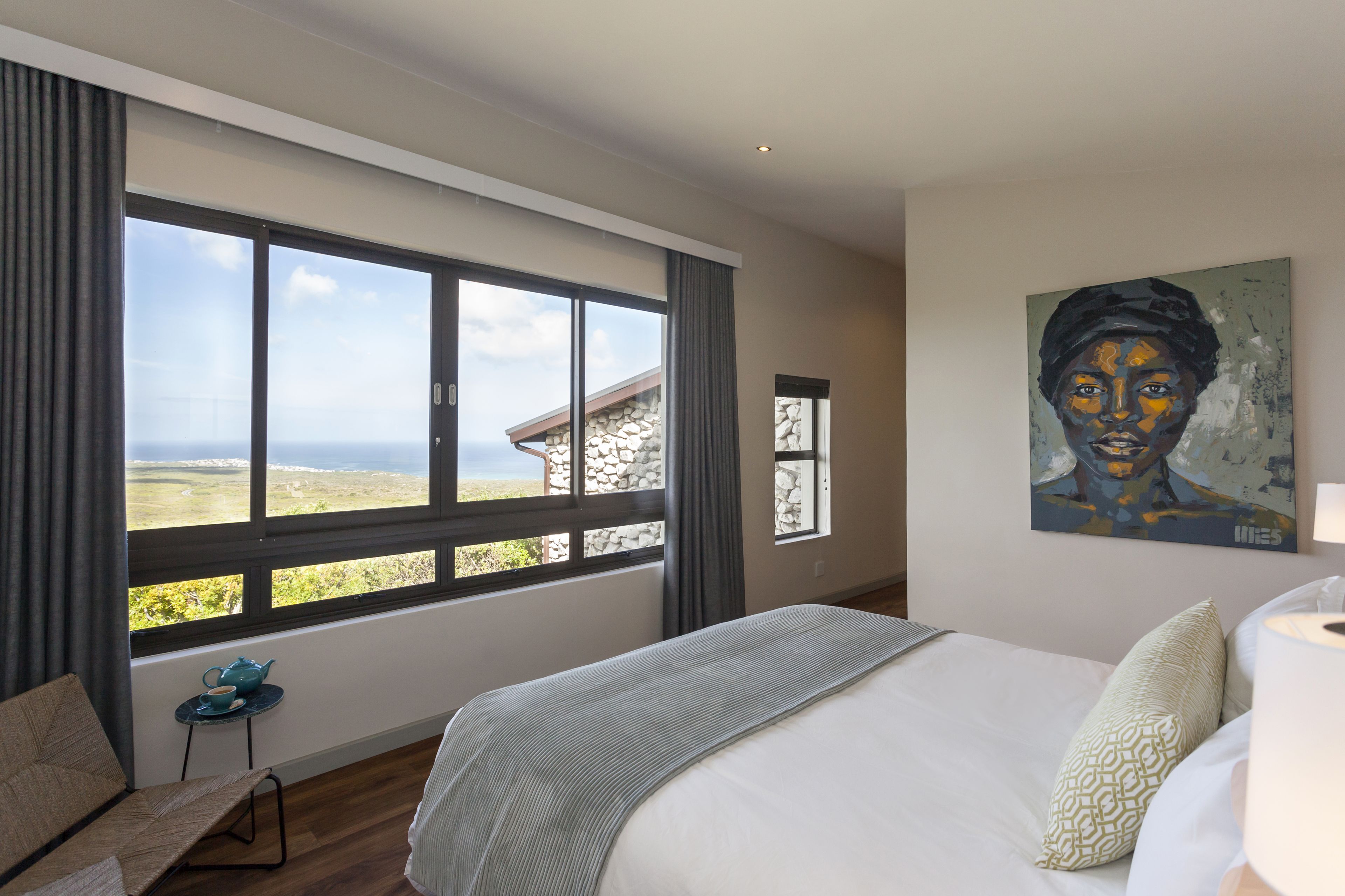 A bedroom with a large window, a bed, a chair, and a painting on the wall.