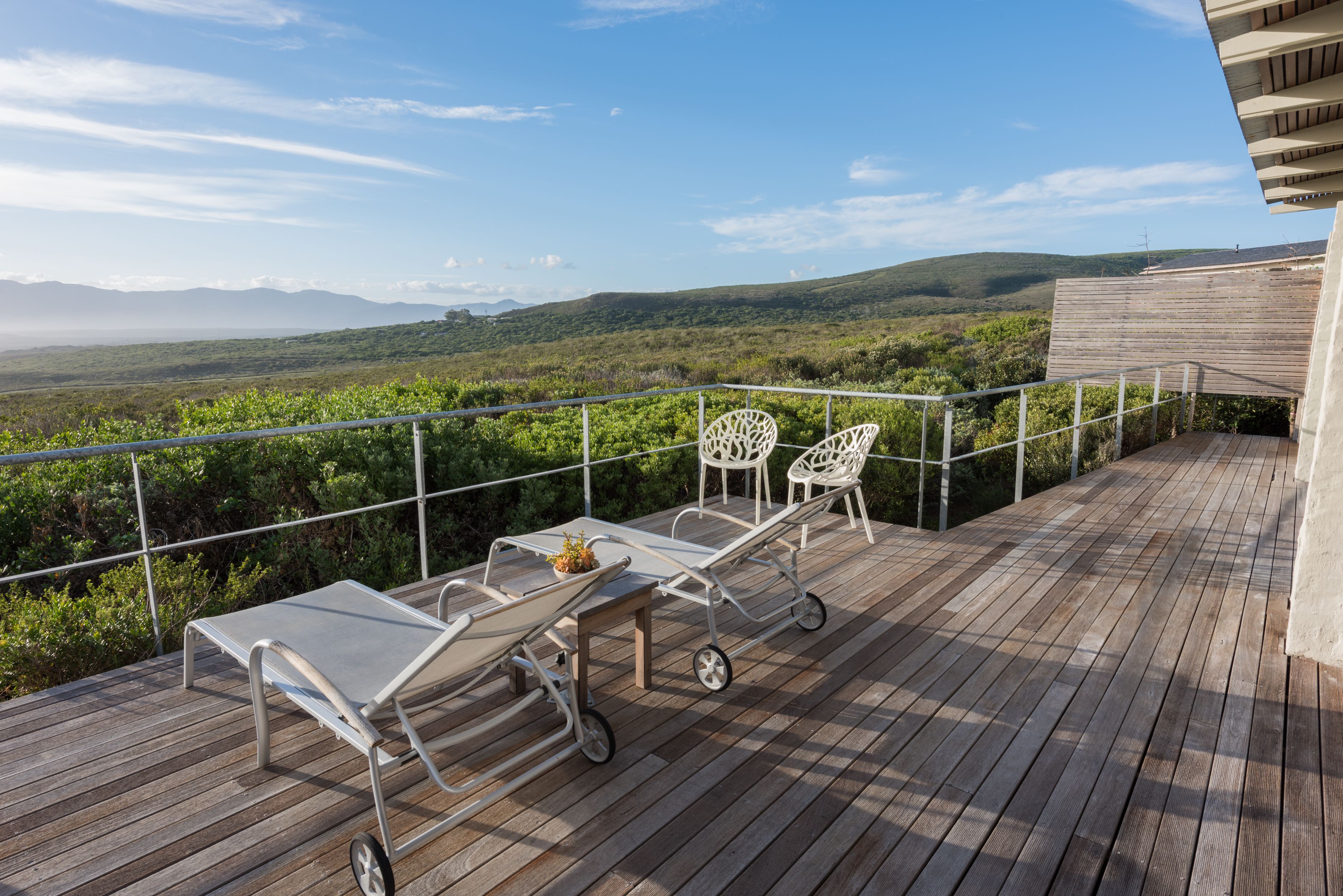 Una terraza de madera con sillas y una mesa de metal, con vistas a un paisaje exuberante y colinas a lo lejos.