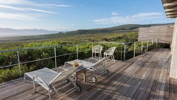 Una terraza de madera con sillas y una mesa de metal, con vistas a un paisaje exuberante y colinas a lo lejos.