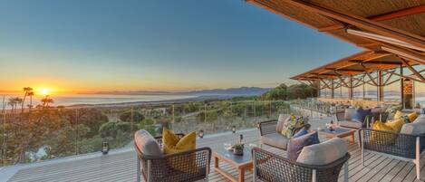 A wooden deck with wicker furniture and a glass balustrade overlooking a scenic landscape during sunset.