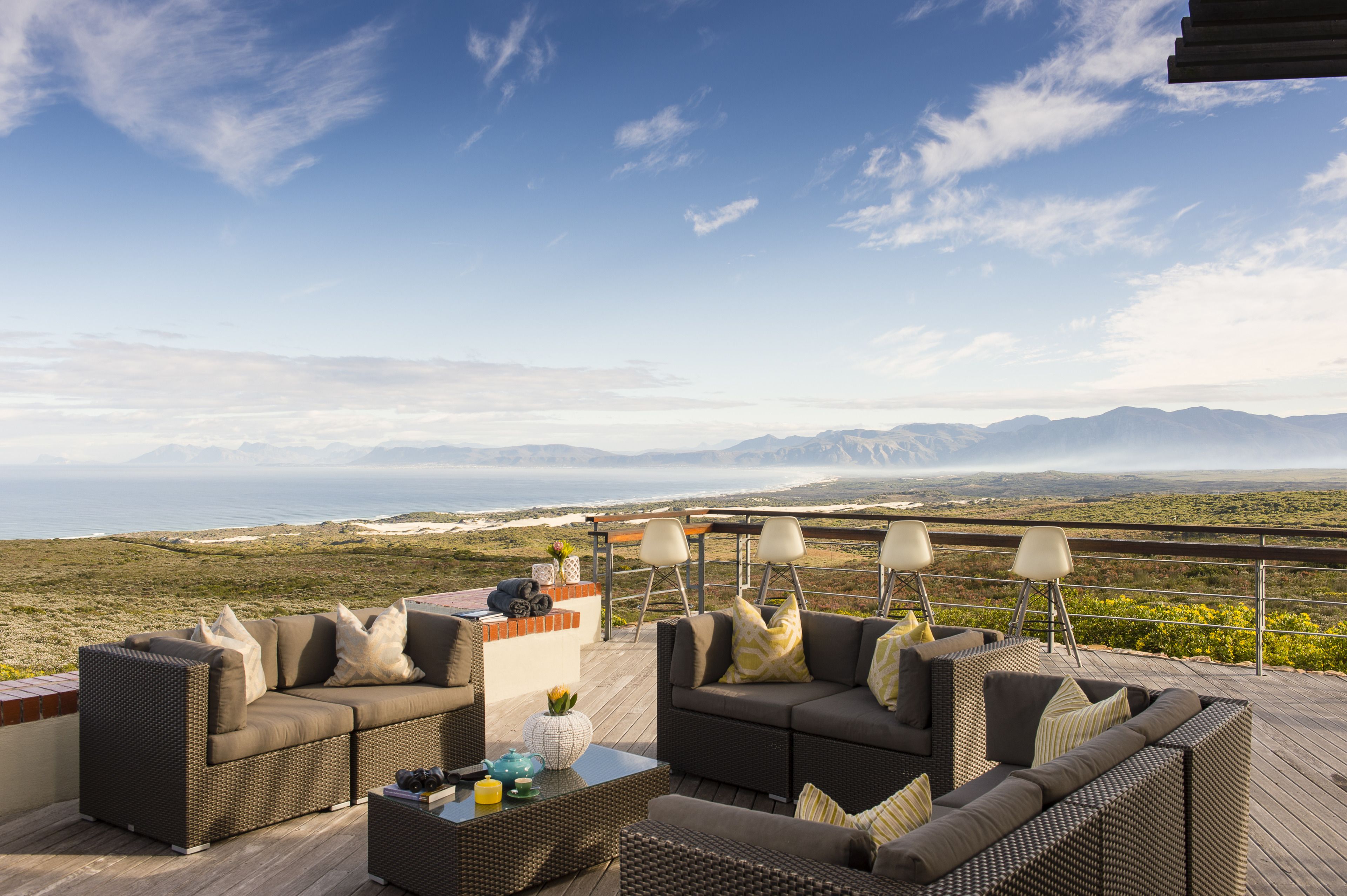 A deck with wicker furniture, a table with a vase and teapot, and a view of the ocean and mountains.