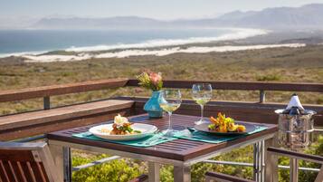 Una mesa dispuesta con platos de comida, copas de vino y un florero con flores, con vista a un paisaje costero.