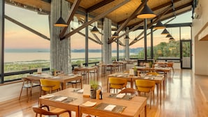 Un restaurante con mesas y sillas de madera, amplias ventanas con vista al mar y las montañas, y lámparas colgantes.