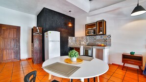 Een compacte keuken met een witte koelkast, een magnetron en een fornuis. Er is een eettafel met stoelen en een potplant.