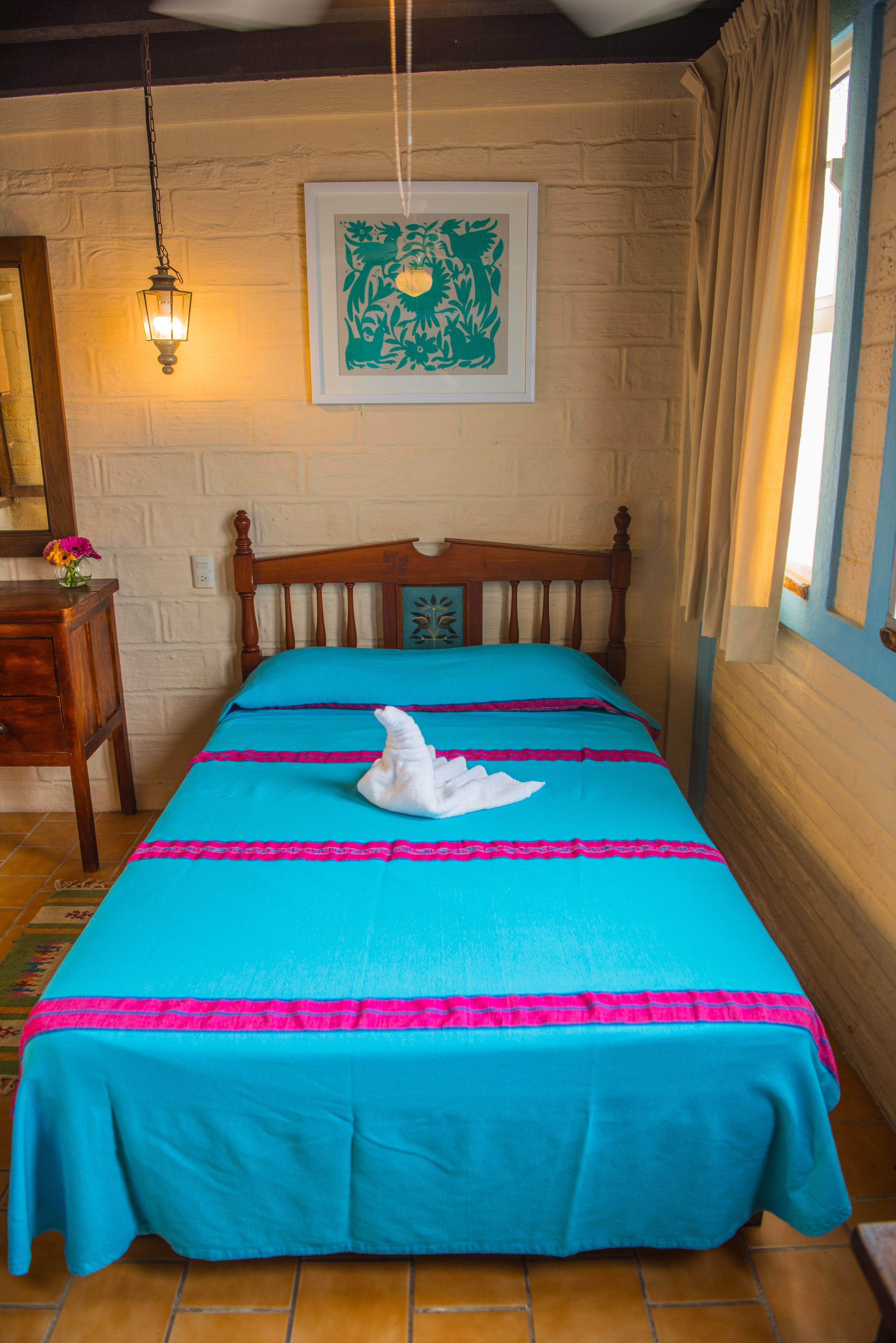 Uma cama com colcha azul e rosa, cabeceira de madeira, luminária fixada na parede e uma obra de arte emoldurada acima da cama.