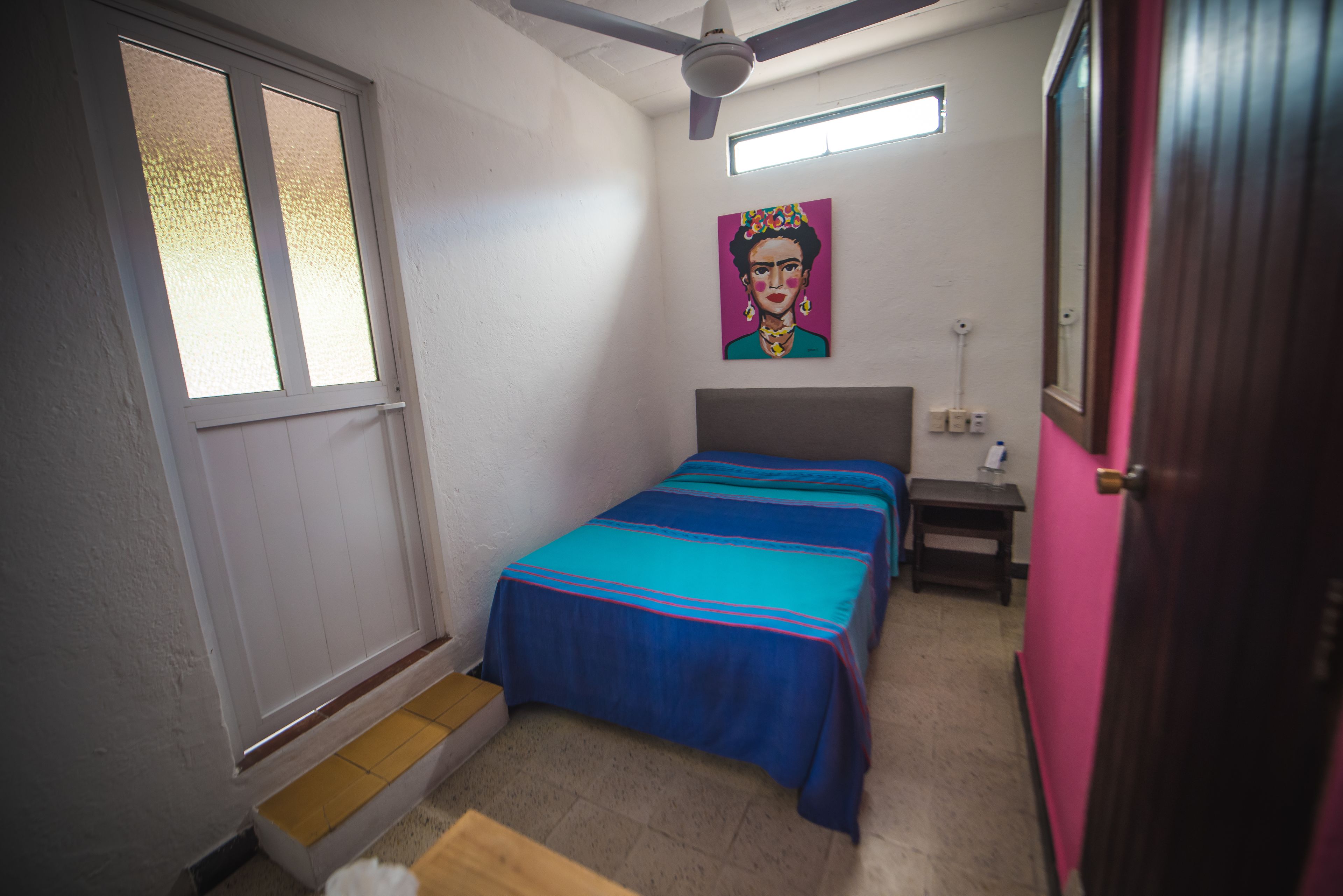 A bedroom with two beds, a ceiling fan, a window, a painting on the wall, and a door.