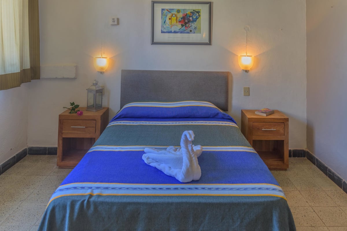 Een opgemaakt bed met een blauw-groen gestreept beddegoed en een witte handdoek erover gevouwen. Twee nachtkastjes, elk met een kleine lamp en een boek. Een ingelijste foto hangt aan de muur boven het bed.
