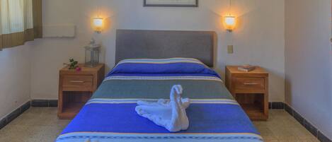 A neatly made bed with a blue and green striped bedspread and a white towel folded on it. There are two bedside tables, each with a small lamp and a book. A framed picture hangs on the wall above the bed.